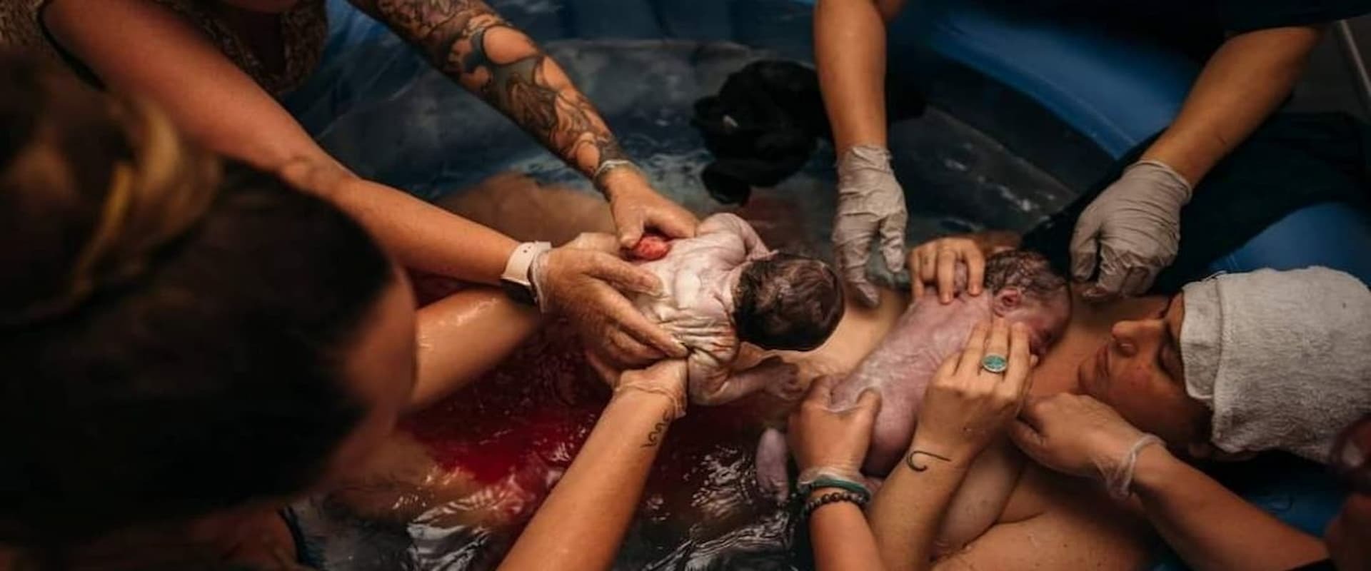 two babies being born water birth with helping hands surrounding the mother in the tub